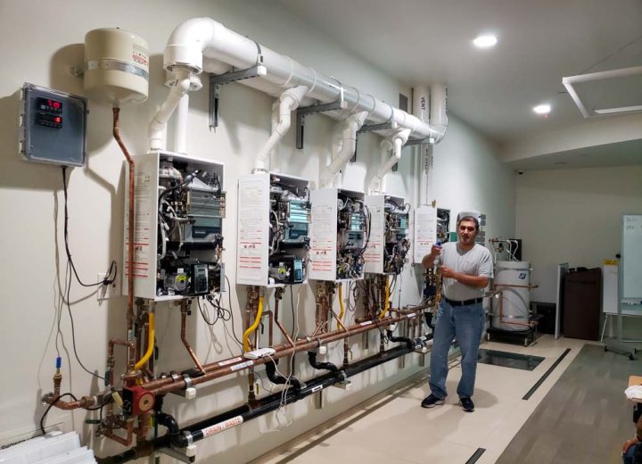 A plumbing instructor standing in a workshop with multiple tankless water heaters mounted for educational purposes.