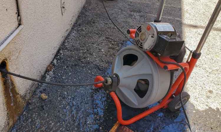Portable electric drain cleaning machine in operation outside, showing cables and water supply connected, ready for heavy-duty use.