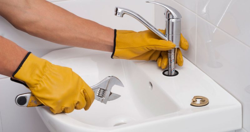 A plumber in yellow gloves installing a modern faucet in a white bathroom sink, showcasing professionalism and cleanliness.