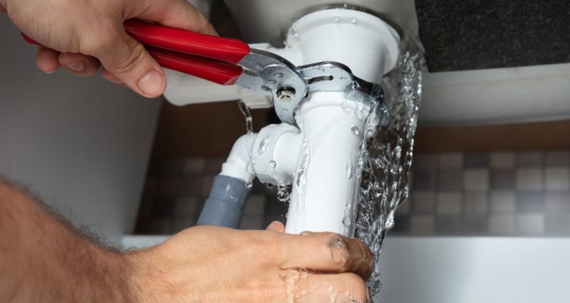 A plumber quickly repairs a leaking white pipe under a sink, water spraying as he works to contain the emergency.
